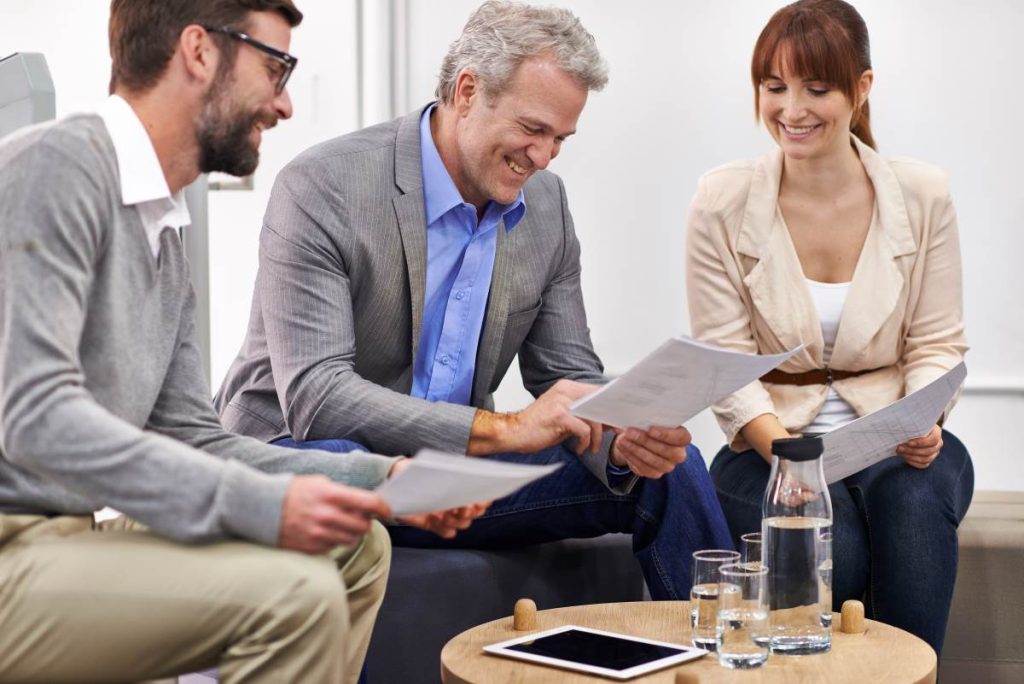 reunião de negócios entre três pessoas, provavelmente em um escritório moderno e bem iluminado. Duas figuras masculinas e uma feminina e